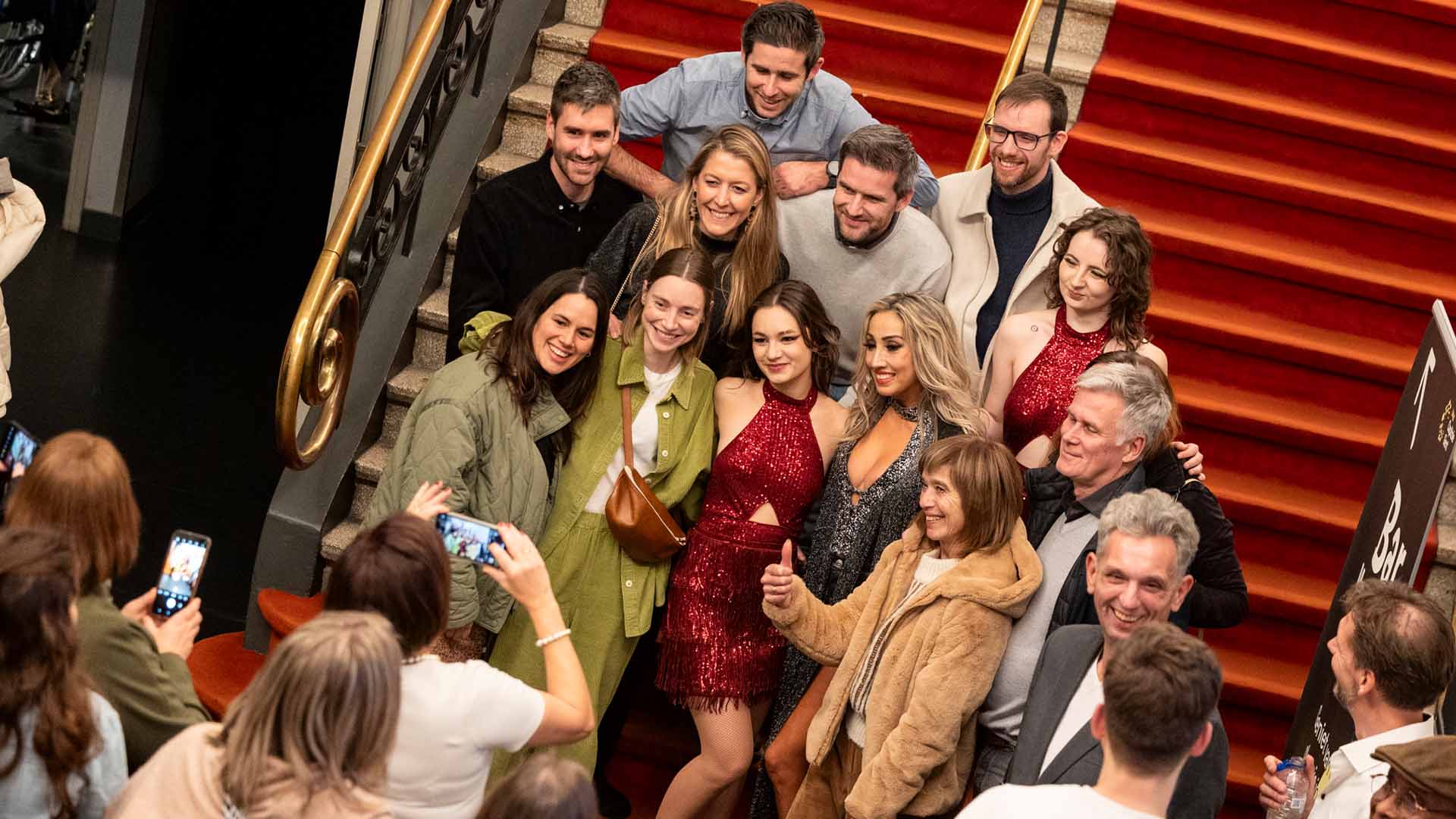 Een groep mensen, sommigen gekleed voor een avondje uit, poseert lachend voor een foto op de rode trap van Capitole Gent, terwijl iemand een selfie maakt.