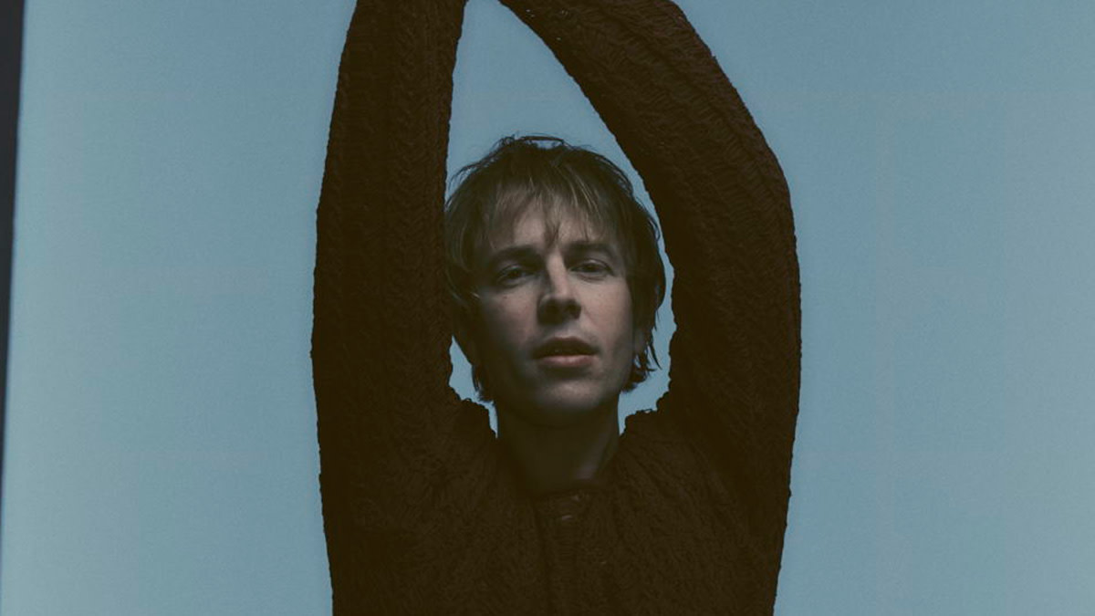 Tom Odell met beide armen in de lucht tegen een blauwe achtergrond kondigt zijn concert op Rock Werchter aan.