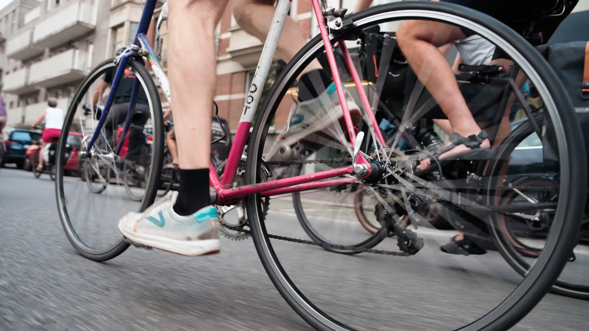 Lage camerahoek van een groep fietsers die samen door de stad rijdt tijdens Oya Vélo. Op de voorgrond zijn de draaiende wielen en trappende benen van verschillende deelnemers zichtbaar.