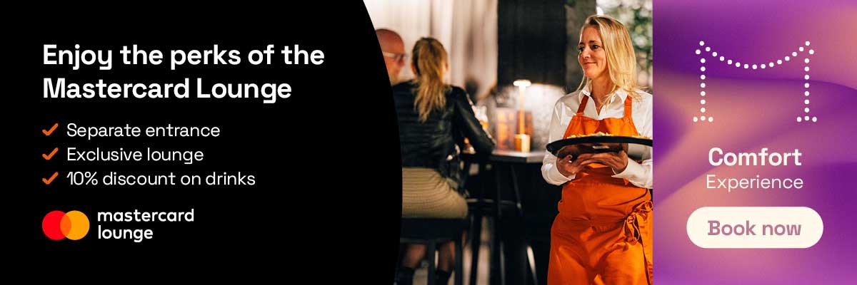Interior of the Mastercard Lounge showing a staff member serving drinks to guests standing in a lounge setting.