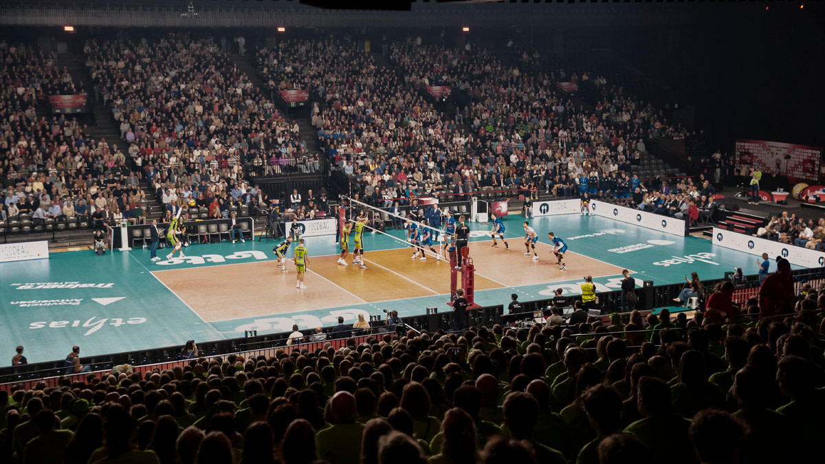 Een volleybalwedstrijd in een uitverkochte AFAS Dome, waar twee teams tegenover elkaar staan aan het net terwijl duizenden fans in de tribunes aandachtig volgen en de actie op het veld versterken met hun aanwezigheid.
