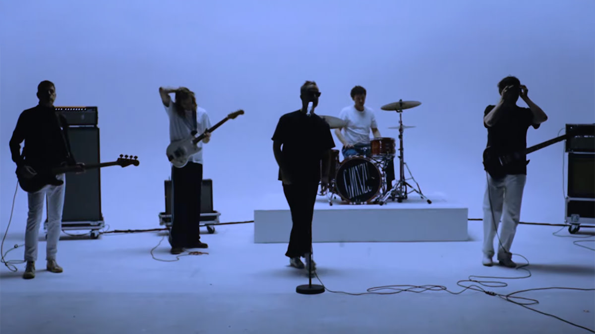 Ghinzu poseert als vijfkoppige band in een strakke witte studio. De bandleden staan verspreid met gitaren en een drumstel, met het Ghinzu-logo zichtbaar op de basdrum.