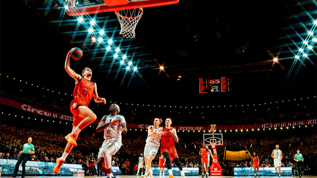 Basketbalspeler van de Antwerp Giants springt naar de ring voor een dunk in een volle Lotto Arena terwijl tegenstanders toekijken en supporters op de achtergrond zichtbaar zijn.