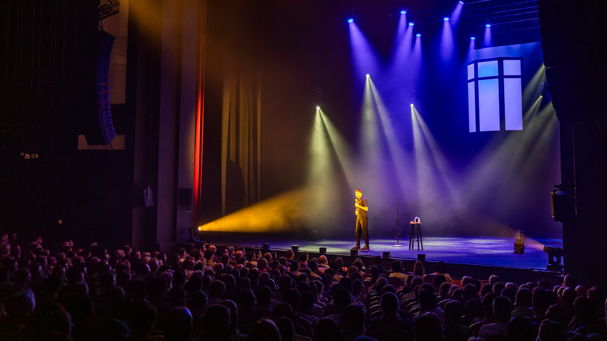 Overzichtsfoto van comedian Mohsin Abbas op een podium, verlicht door een gele spotlight. Hij staat voor een groot, donker publiek in een gevulde theaterzaal met blauw-paarse podiumverlichting.