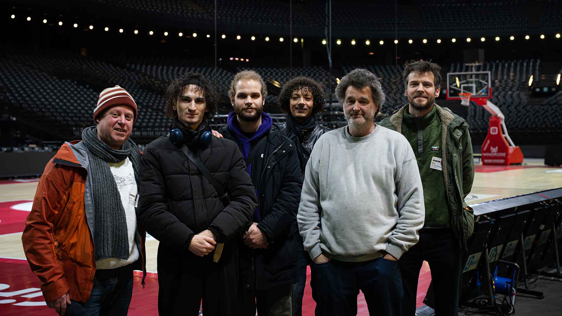 Een crewlid van de AFAS Dome en de Lotto Arena staat samen met enkele jongeren van Elegast Den Dam in de AFAS Dome, met tribunes en sportinfrastructuur op de achtergrond tijdens een kennismakingsmoment achter de schermen.