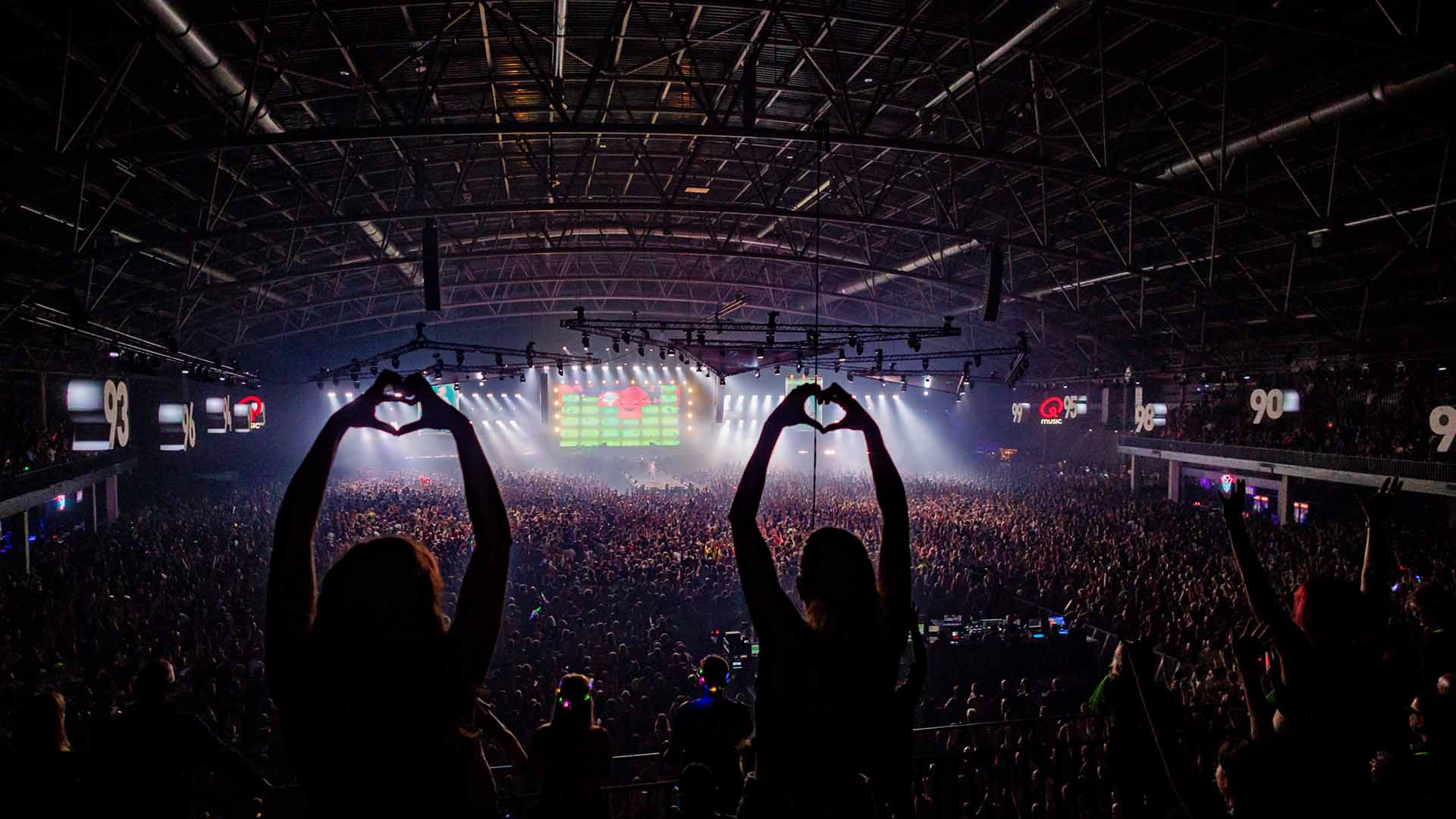Twee vrouwen kijken uit op een volle dansvloer en vormen hartjes met hun handen op I Love The 90s.