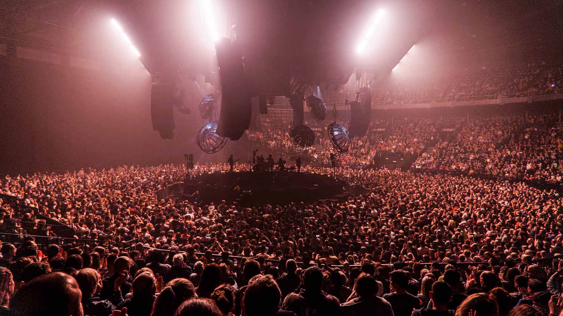 Au centre de la salle se trouve une scène ronde. La scène est plongée dans le noir, seuls les silhouettes des artistes sont visibles. Des fans entourent la scène et les gradins sont pleins.
