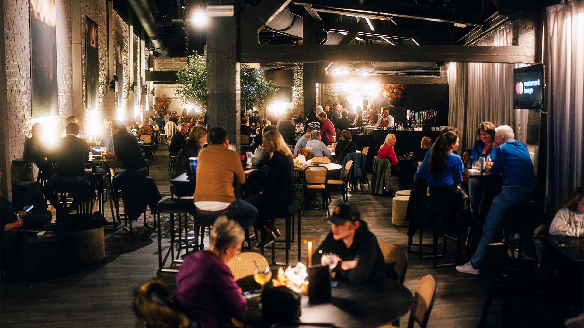 In the Mastercard Lounge, people are sitting at various tables chatting. The ambient lighting creates a warm and cosy atmosphere.