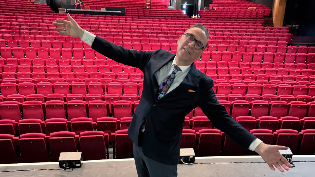 Peter Douglas, internationaal gerenommeerd vertolker van Frank Sinatra, poseert met gespreide armen op het podium van Stadsschouwburg Antwerpen. Op de achtergrond vullen rijen rode theaterstoelen de zaal.