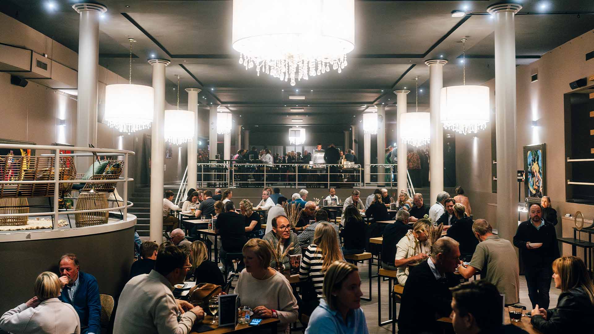 In de VIP Village staan verschillende hoge tafeltjes opgesteld. Mensen eten, drinken en praten aan de tafeltjes. Aan het plafond hangen verschillende felverlichte kroonluchters.