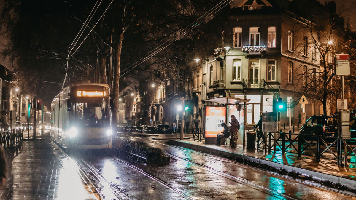 Tram rijdt ’s avonds door een regenachtige straat in Brussel met wachtende reizigers aan een verlichte halte.