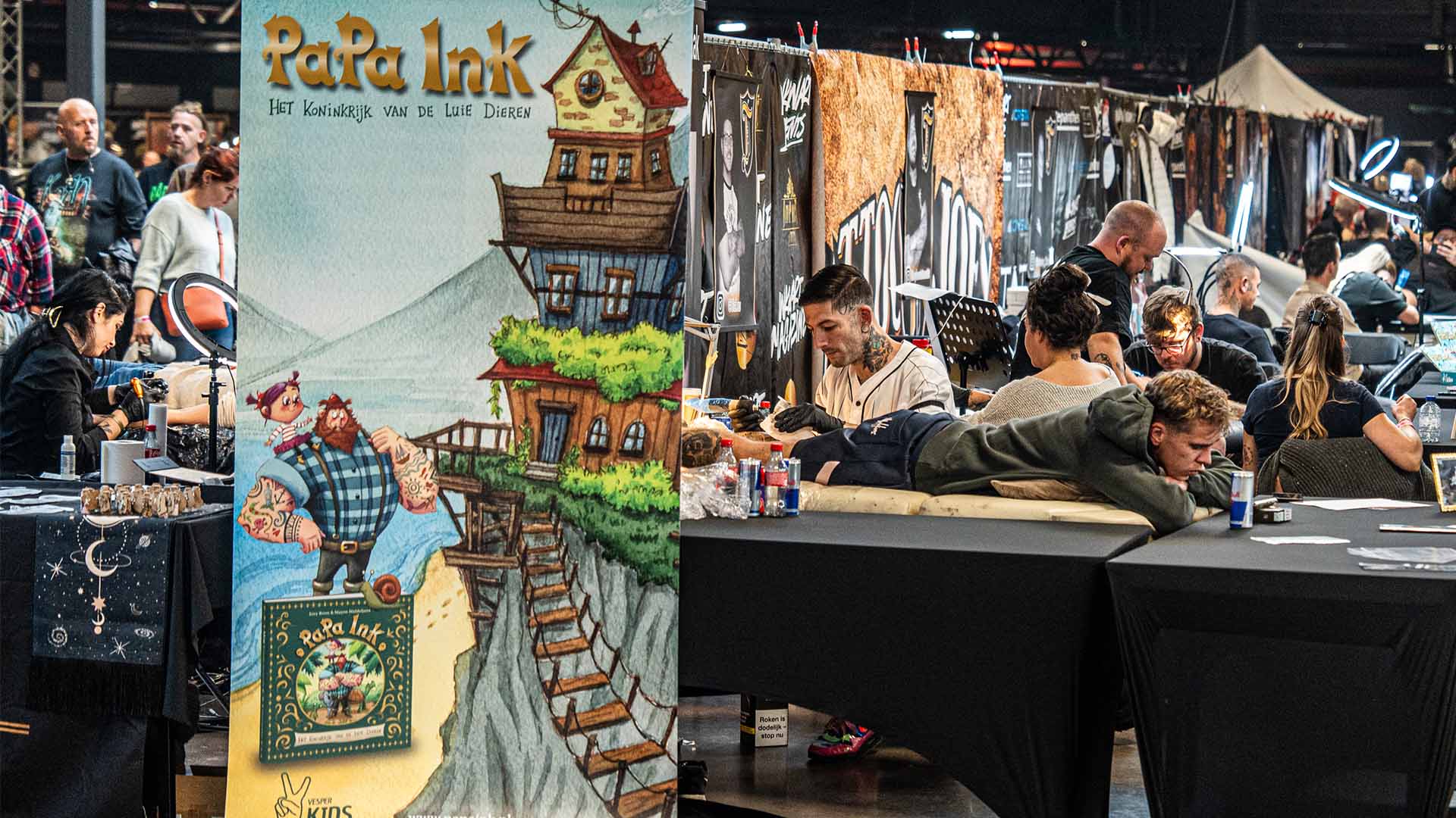 Verschillende tattootafels staan naast elkaar opgesteld en enkele mensen worden getatoëeerd. Op de voorgrond is een affiche te zien van het boek 'Papa Ink: het koninkrijk van de luie dieren'. 