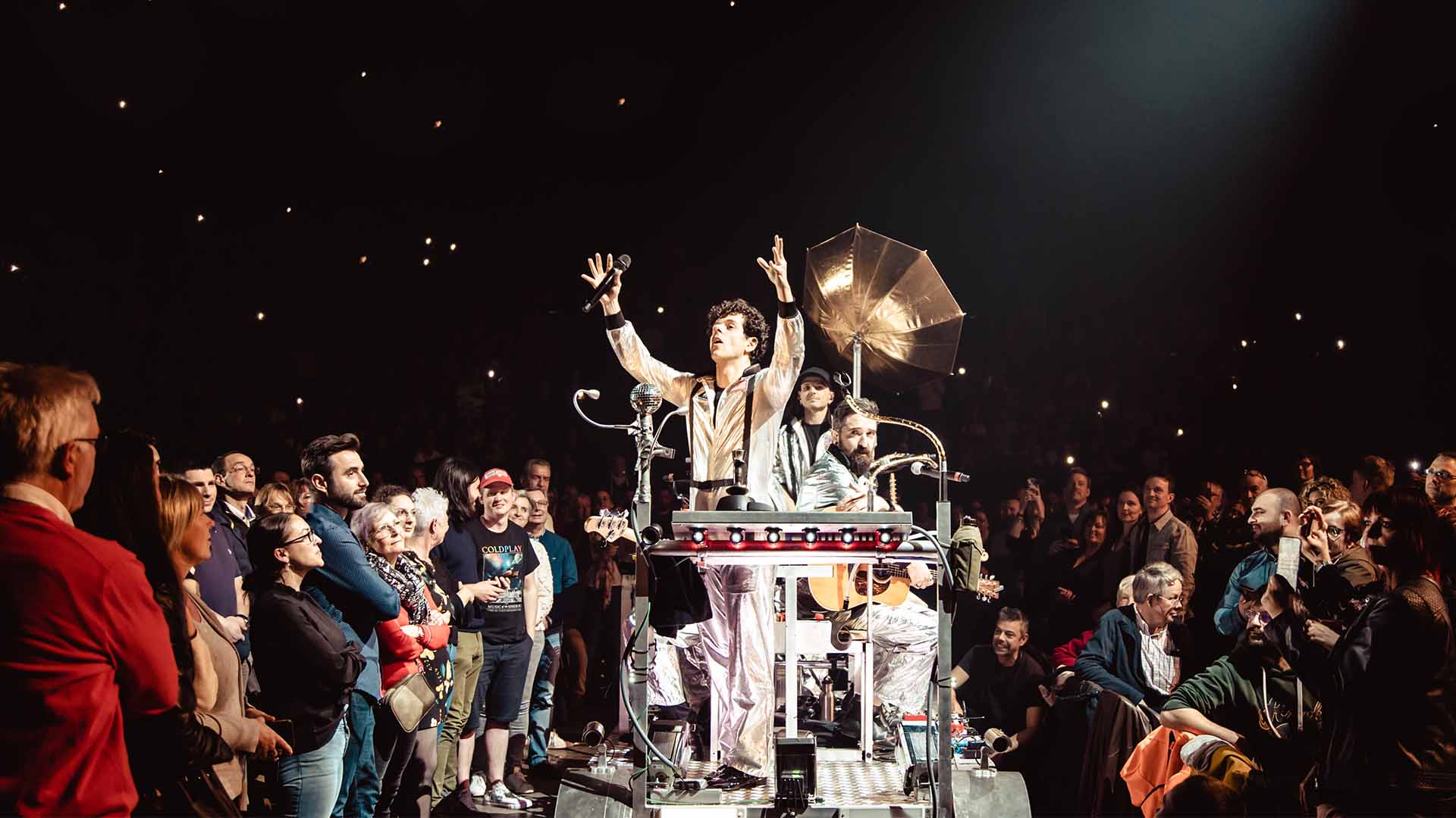 Michael Gregorio and two musicians are moving through the venue on a mobile platform. They are wearing spacesuits and are surrounded by the audience.