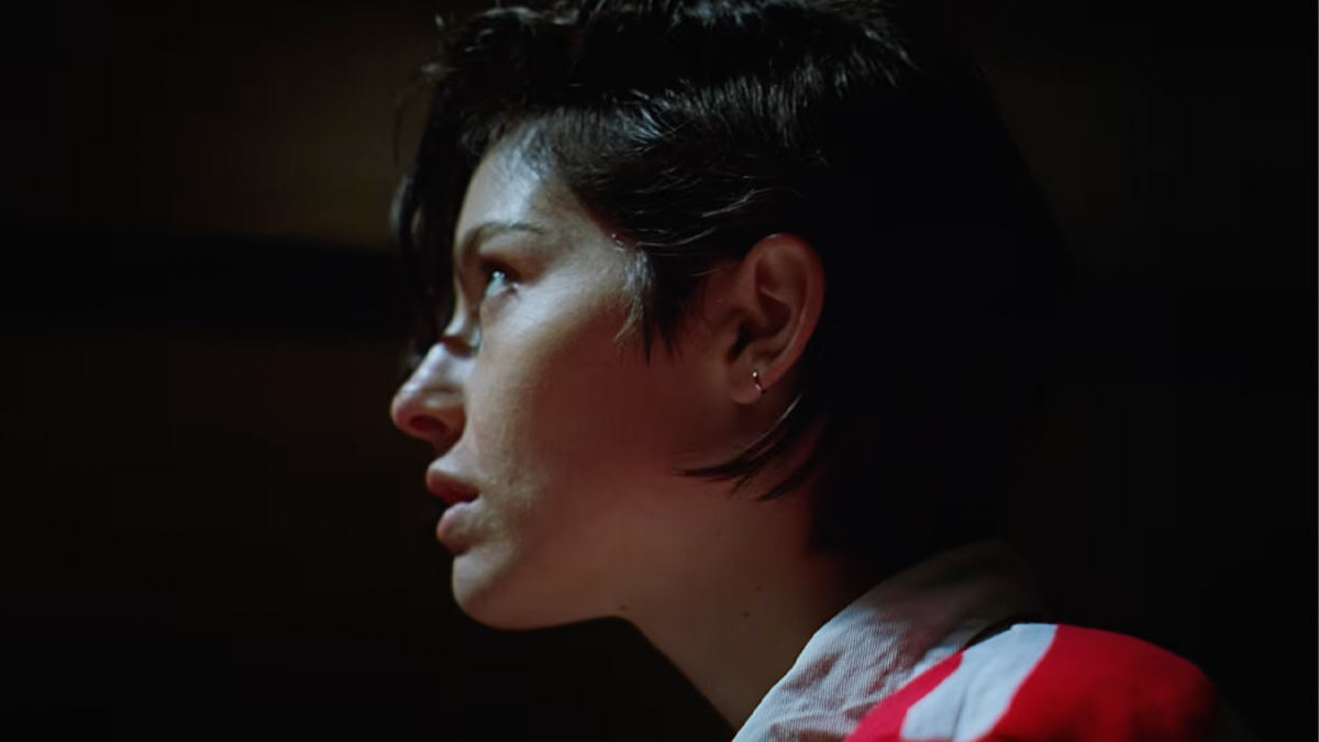Close-up van Gracie Abrams in zijprofiel met kort donker haar, sfeervol belicht in een donkere ruimte met warme rode en bruine tinten.