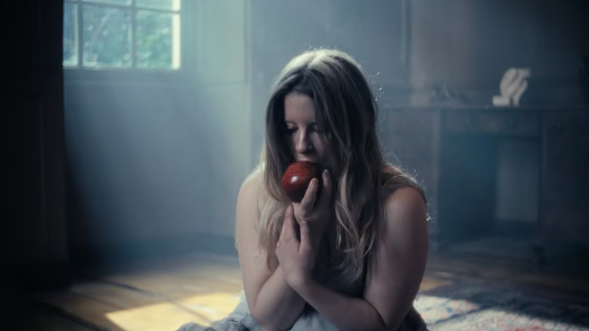 Paris Paloma zit op de vloer van een schemerige kamer en houdt een rode appel dicht tegen haar gezicht, terwijl zacht daglicht door een raam naar binnen valt.