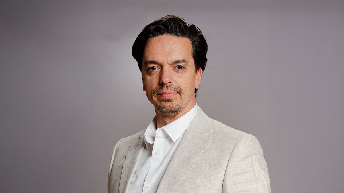 Portret van Nordin De Moor tegen een neutrale grijze achtergrond. Hij kijkt recht in de camera en draagt een licht beige blazer over een wit hemd, met een verzorgde baard en donker, naar achter gekamd haar.