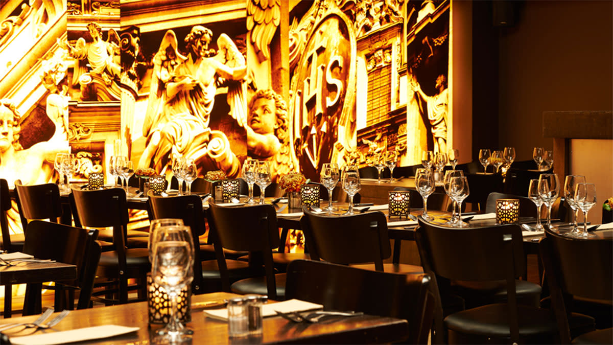 Gezellige brasserie met sfeervol gedekte tafels en warme verlichting, met op de achtergrond een opvallende goudkleurige muurschildering met klassieke beelden en religieuze elementen in Brasserie Appelmans in Antwerpen.