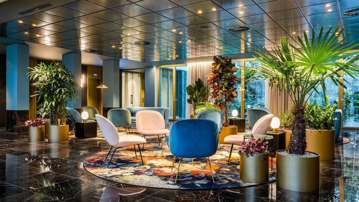 Lobby élégante du NH Collection Antwerp Centre avec fauteuils colorés, éclairage design, plantes tropicales et espace lounge moderne autour d’un tapis rond.