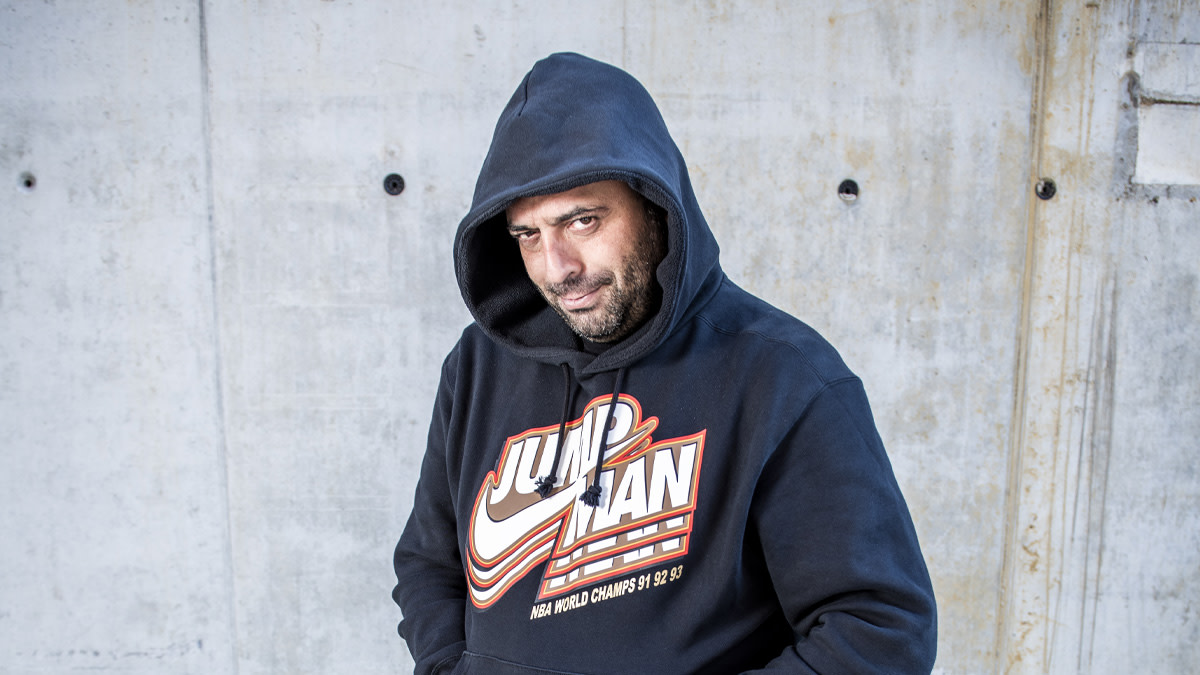 Comedian Erhan Demirci in een donker blauwe hoodie tegen een betonnen wand. 