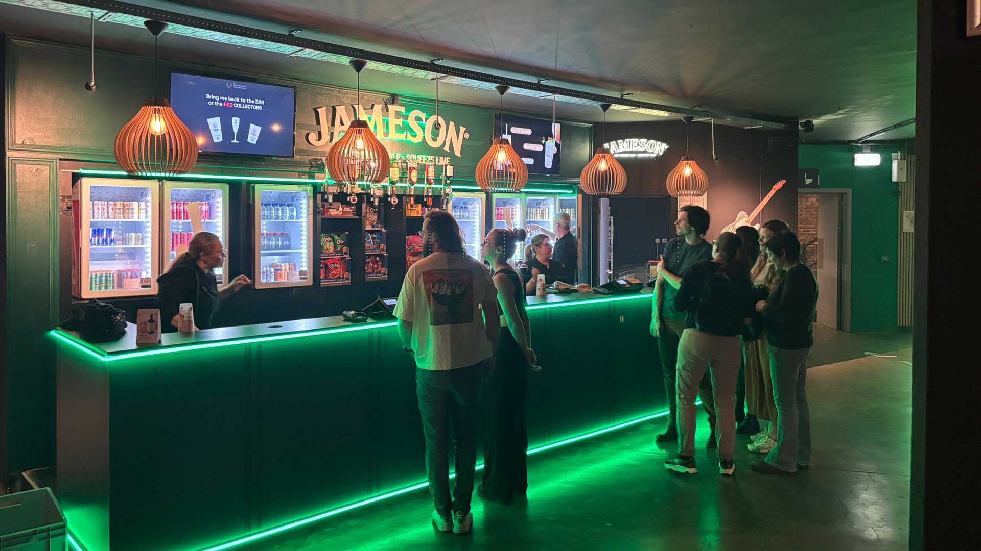 Bar Jameson à Forest National à Bruxelles, avec un éclairage d’ambiance vert, un comptoir illuminé et des réfrigérateurs derrière le bar. Les visiteurs commandent des boissons dans la salle de concert couverte de Forest National.