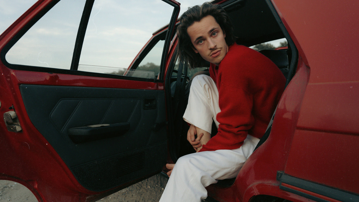 Iskander Moon in a red sweater and white trousers sitting in the backseat of a red car with the door open, looking at the camera.