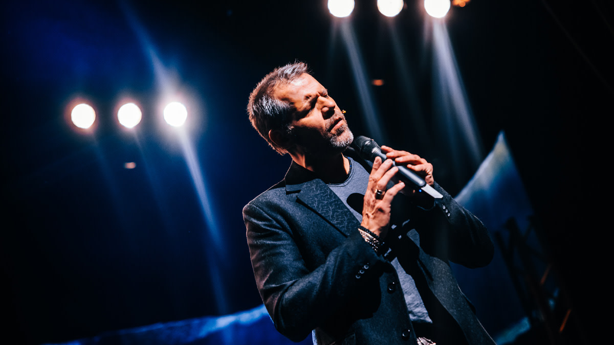 Musicalster René van Kooten zingt met gesloten ogen en veel emotie op het podium, terwijl hij een microfoon vasthoudt onder theaterlichten.