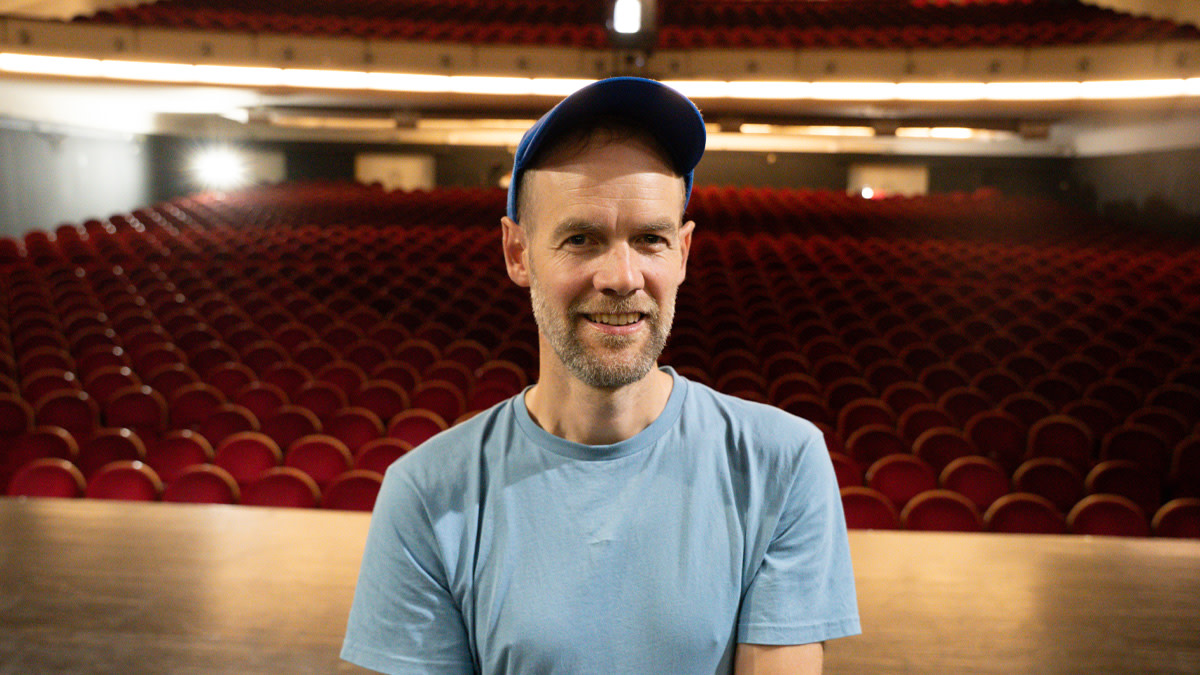Wannes Cappelle van Het Zesde Metaal staat op het toneel en kijkt lachend in de camera, met een lege theaterzaal vol rode stoelen achter hem.