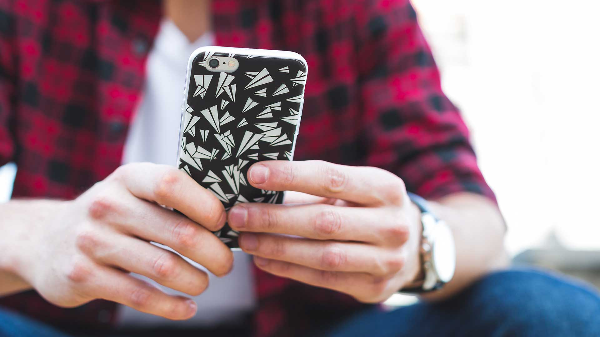 Mains tenant un smartphone avec une coque à motifs noir et blanc. La personne porte une chemise à carreaux rouge et noir et une montre. L’accent est mis sur l’appareil et son utilisation.