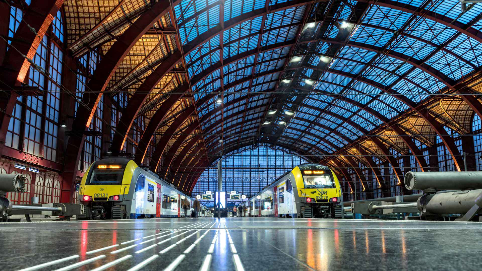 Intérieur de la gare centrale d’Anvers avec deux trains à des quais opposés, sous une grande verrière avec une structure métallique et des reflets lumineux sur le sol.