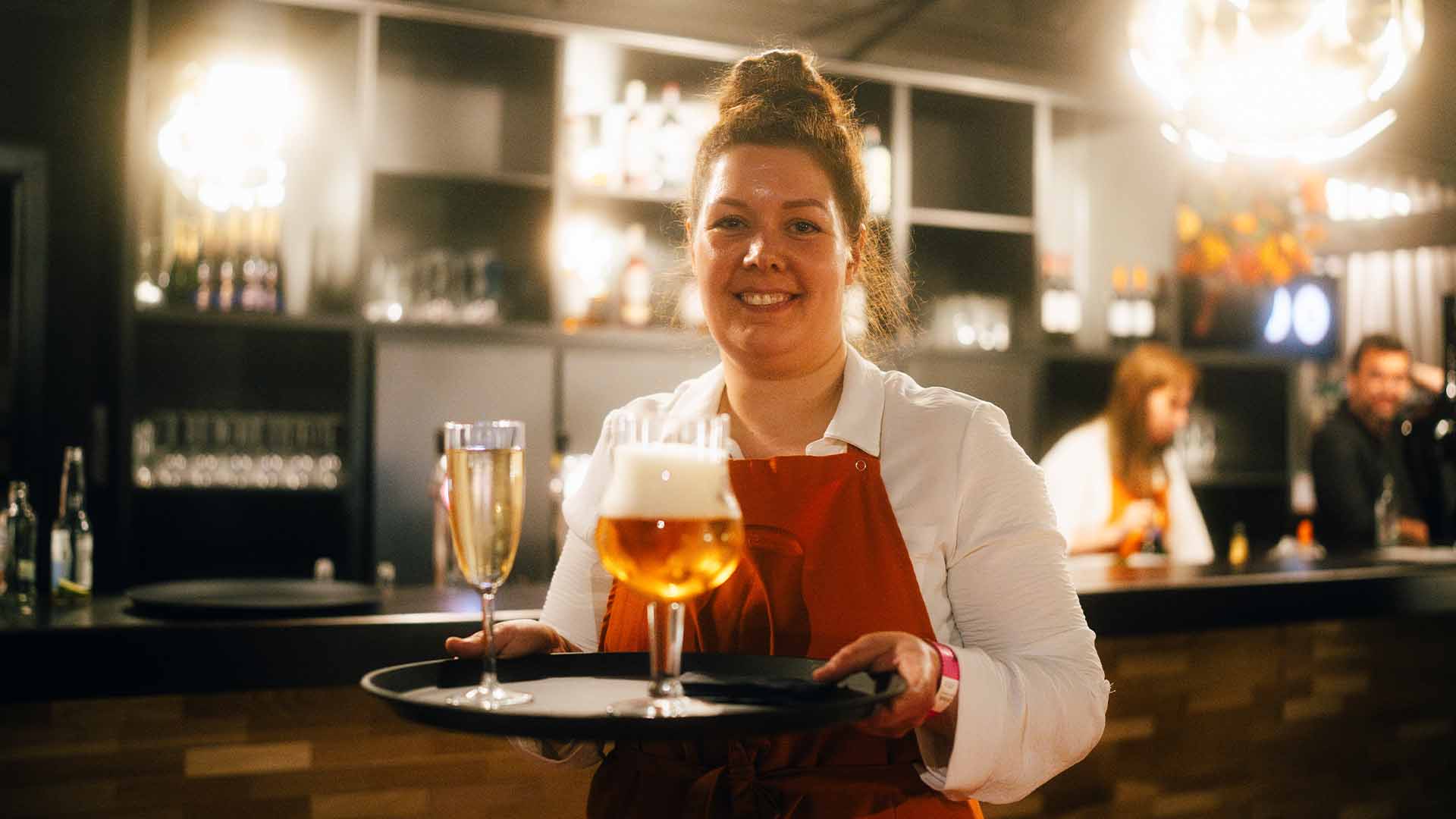 Een glimlachende serveerster houdt een dienblad vast waar een glas champagne en een glas bier op staan.