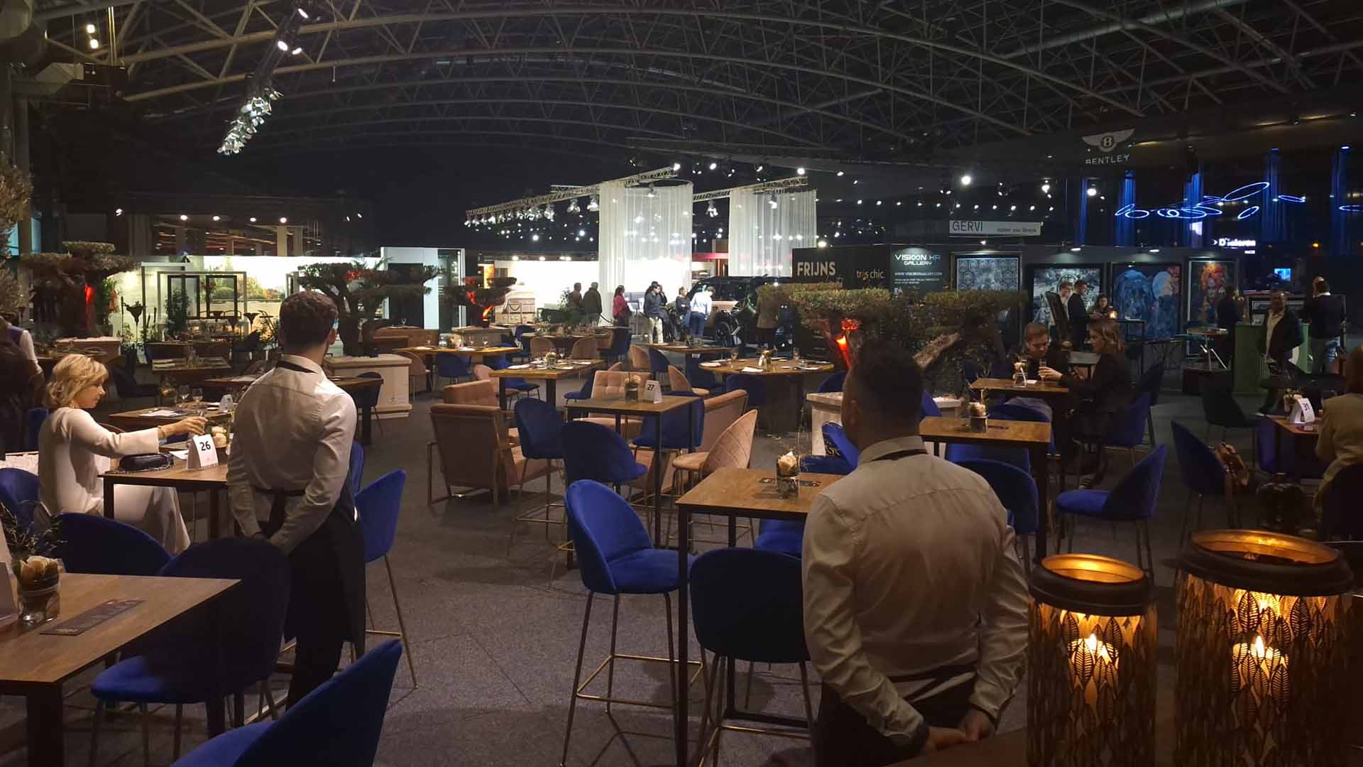 Op de luxueuze lifestylebeurs 'Très Chic' is een hoekje ingericht waar mensen iets kunnen eten en drinken. Er staan verschillende hoge tafeltjes opgesteld en enkele obers staan klaar om bezoekers te bedienen.