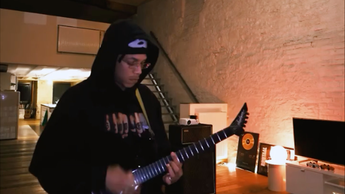 Osayuki Asemota met hoodie en muts speelt elektrische gitaar in een warme, sfeervol verlichte repetitie- of studioruimte, met versterkers en muziekapparatuur op de achtergrond.