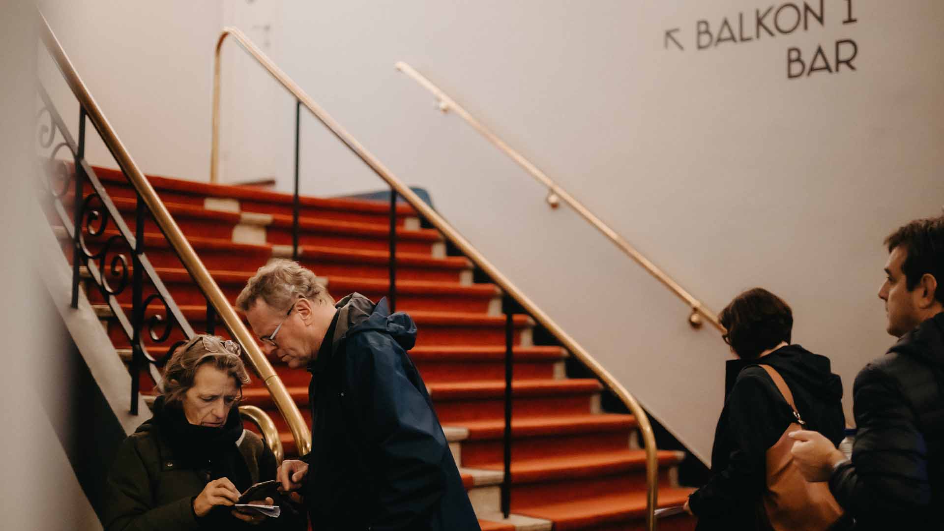 Bezoekers van Capitole Gent staan bij de rode trap, met een bord dat de weg wijst naar 'BALKON 1' en 'BAR'.