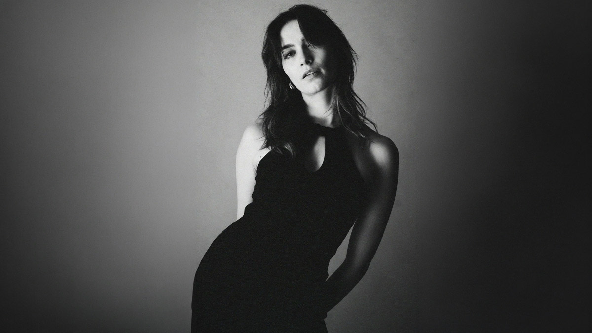 Zwart-wit portret van een vrouw in een elegante, mouwloze zwarte jurk, licht voorovergebogen en recht in de camera kijkend tegen een neutrale achtergrond.