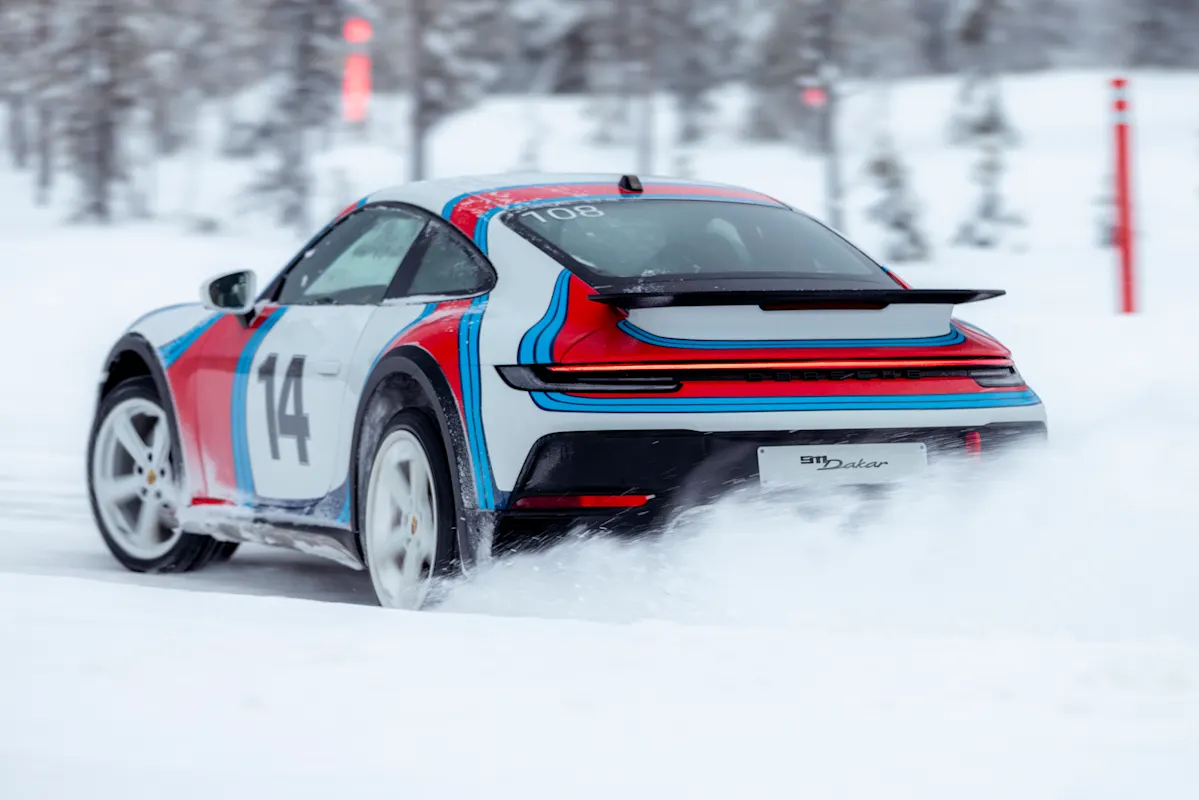 Porsche sports car slides sideways on snowy road, snow flying, trees in background.