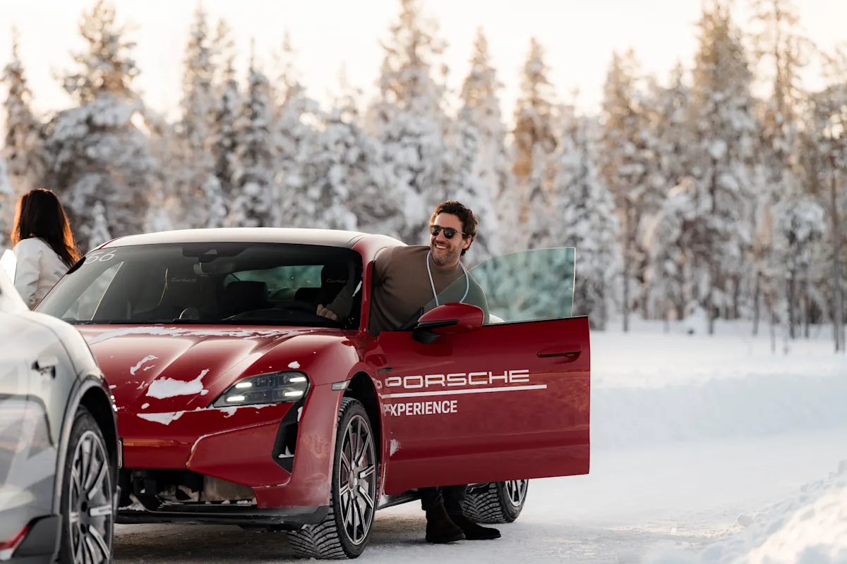 Man smiling stepping out of red Porsche on snowy road, surrounded by snow-covered trees.