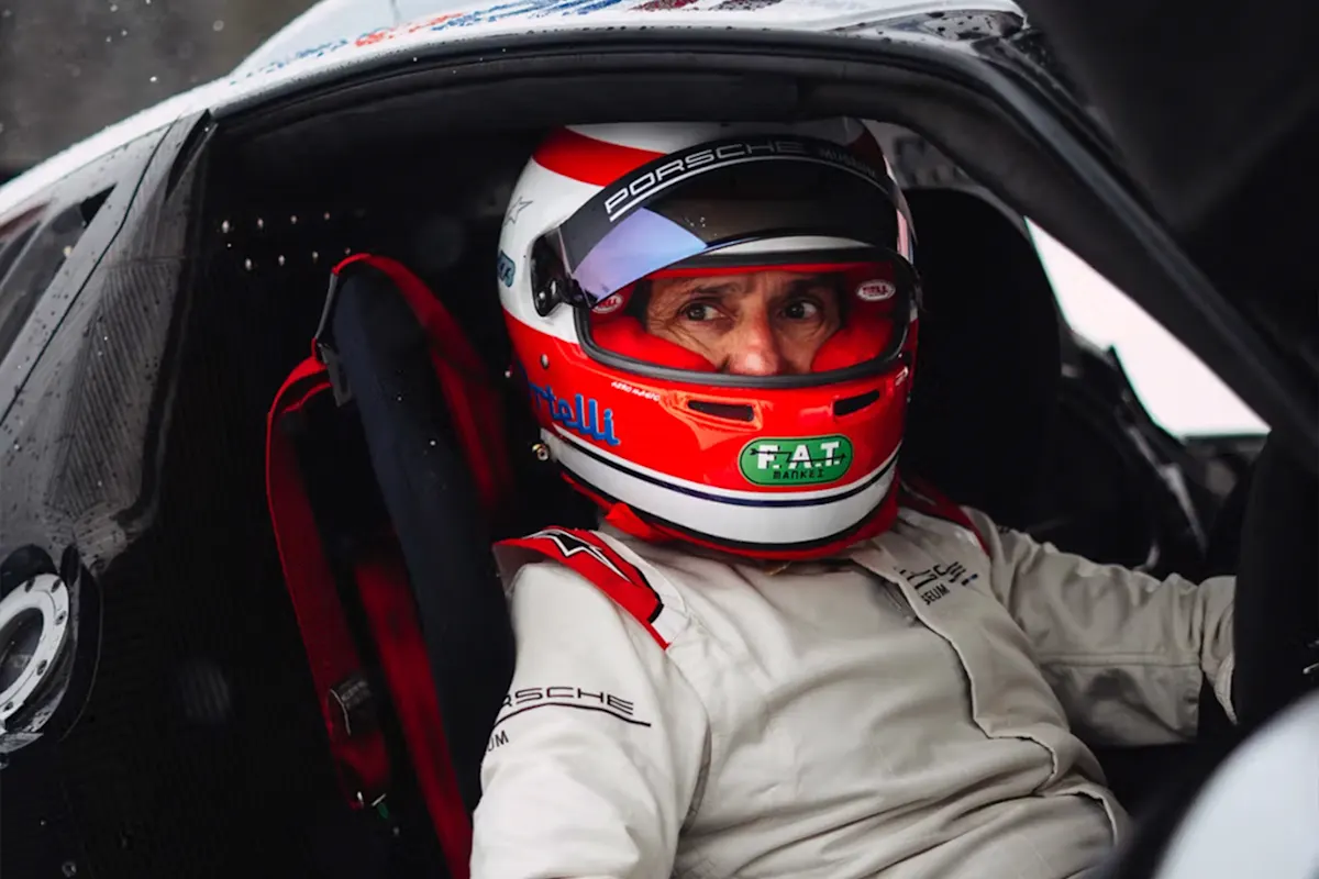 Person in Porsche race car with red and white helmet and suit, seated inside, looking sideways.