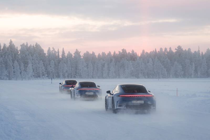 Three Porsche cars on a snowy road with trees and a pastel sunset.