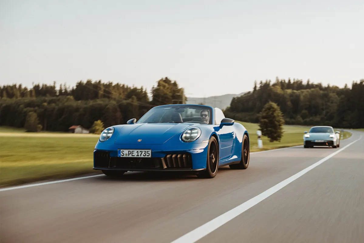 Blauer Porsche fährt auf Landstraße mit Bäumen und Auto dahinter.