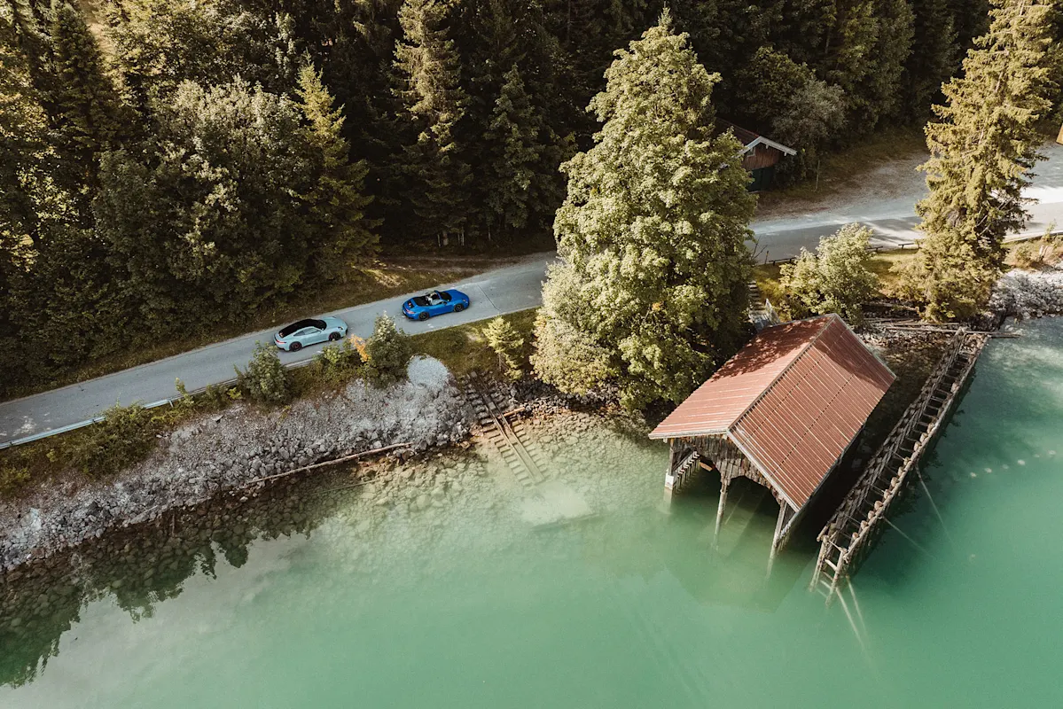 Luftaufnahme von zwei Autos neben einem Bootshaus am See, umgeben von Bäumen.