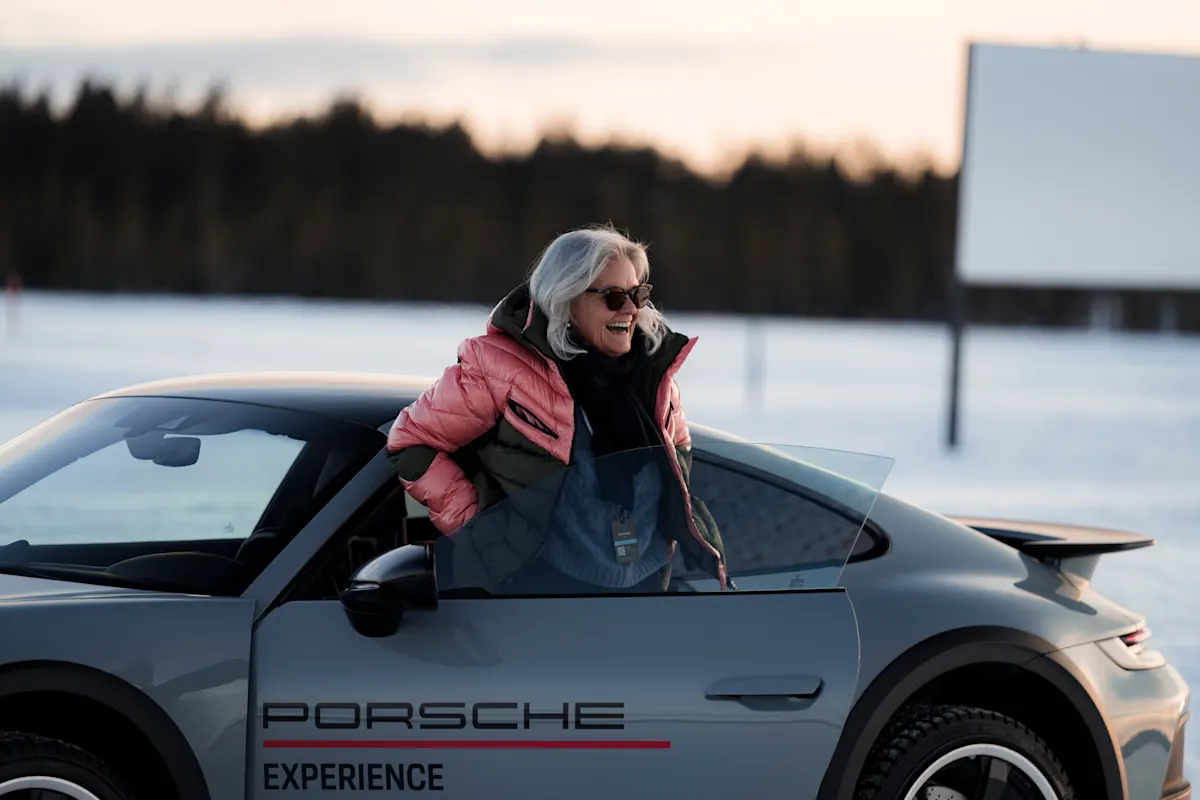 Smiling woman in pink jacket by gray Porsche car in snowy setting.