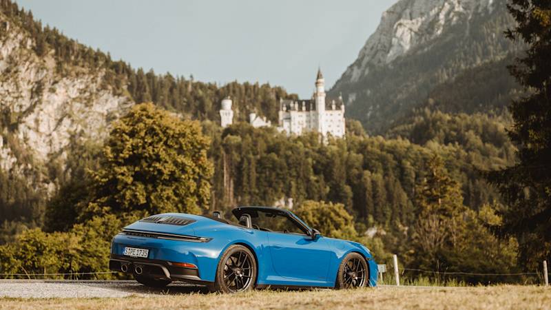Blauer Porsche auf Wiese vor Schloss und Bergen an sonnigem Tag.