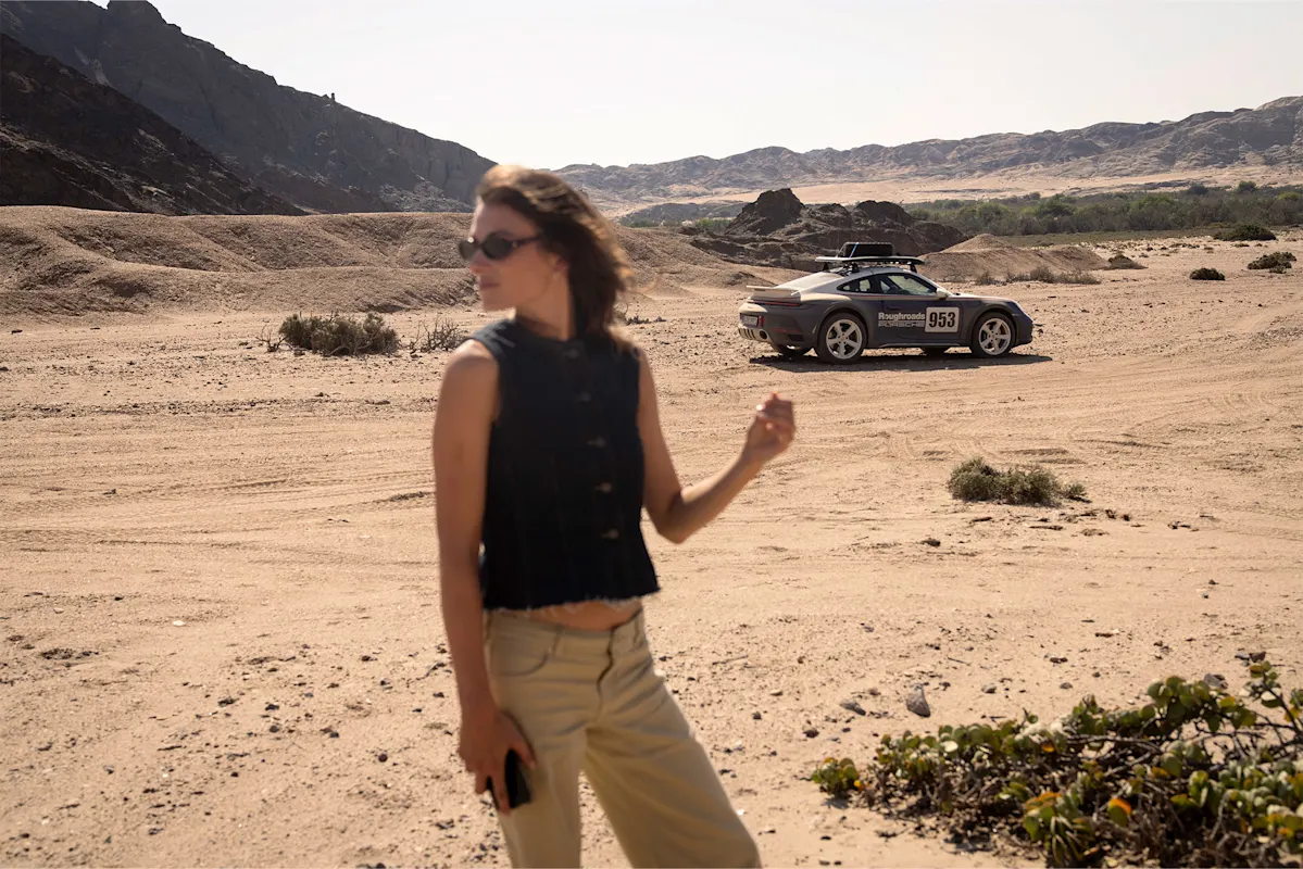 Frau in Wüste mit unscharfem Porsche im Hintergrund.