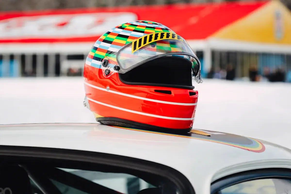 Colorful racing helmet on white Porsche car roof, with blurred red/yellow building in background.