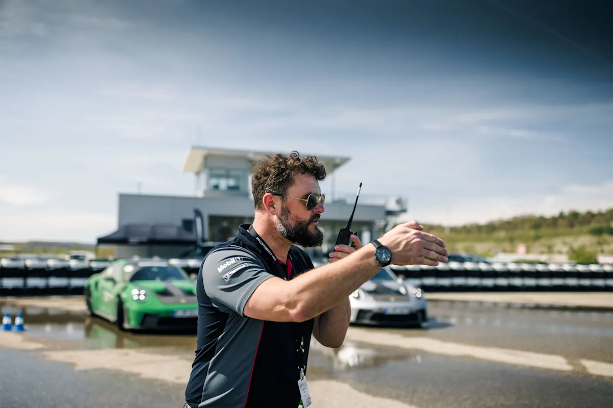 Person with walkie-talkie on Porsche track, with cars and building in background.