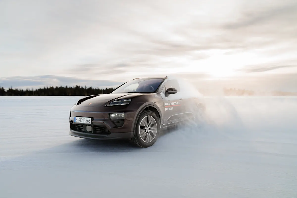 Black Porsche SUV speeds through snowy landscape, forest and clouds in background.