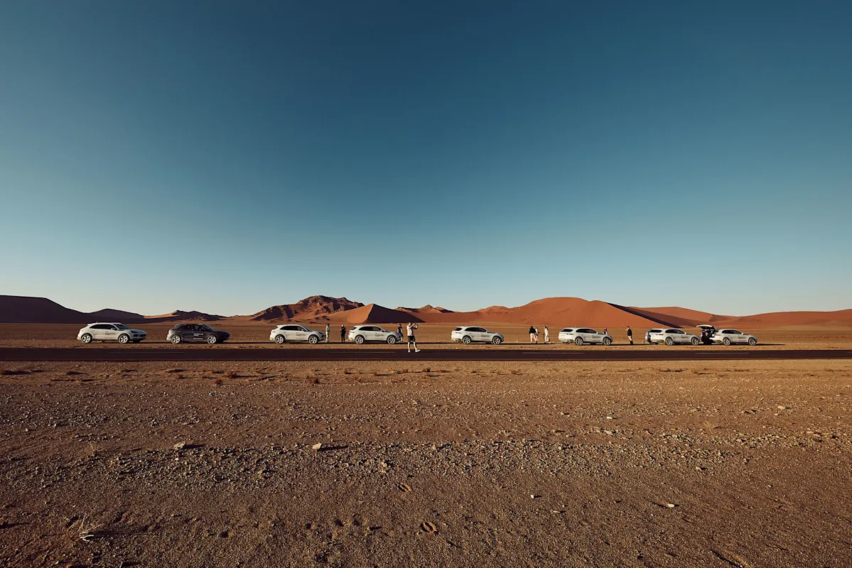 Weiße Porsche SUVs in Wüste, mit Zuschauern und roten Dünen dahinter.