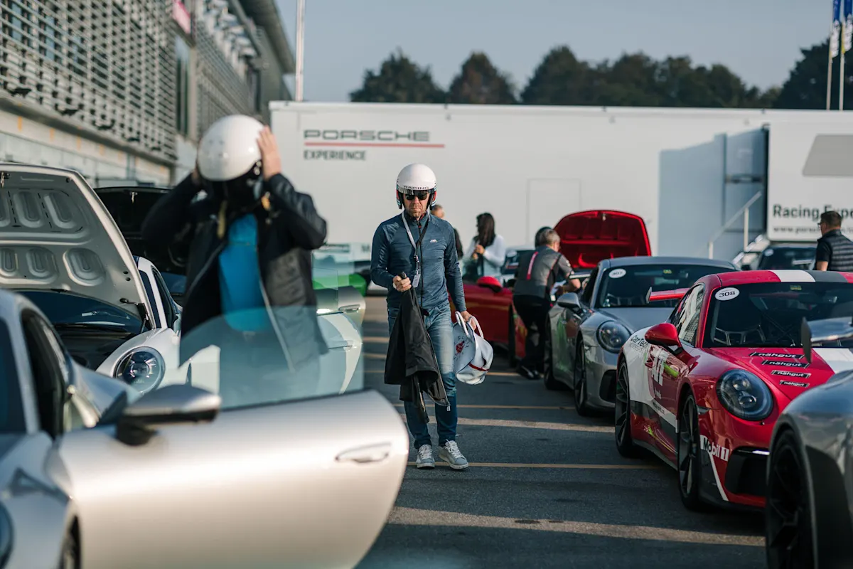 Participants prepare for a track session in the pit lane, standing between several parked Porsche vehicles.