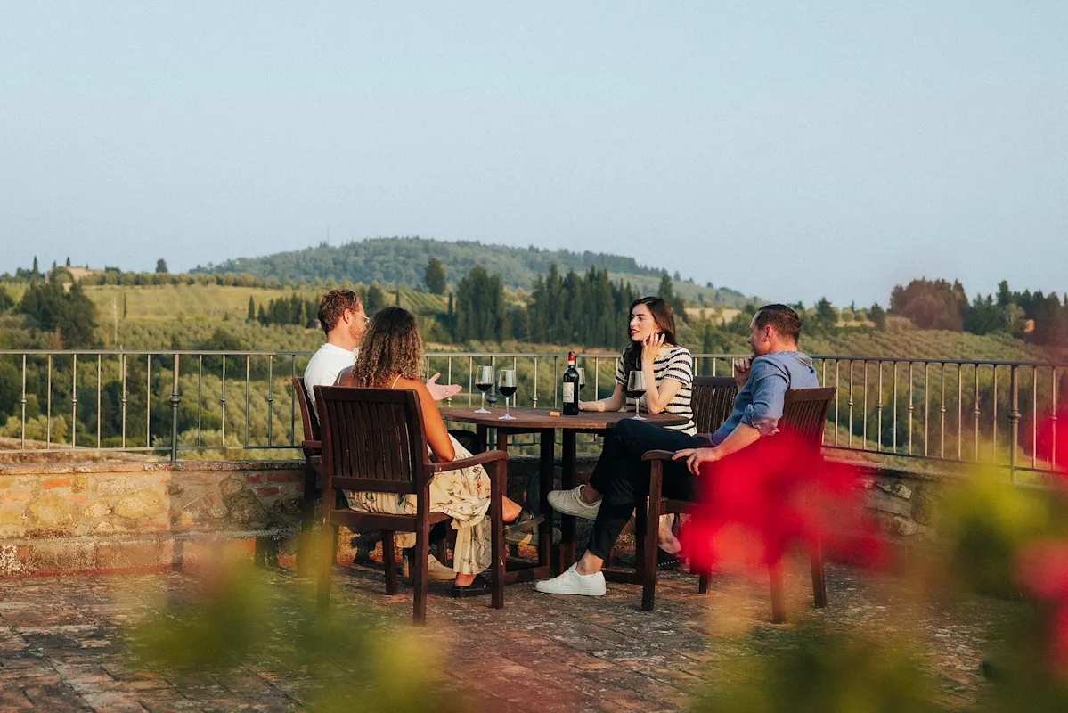 Vier Personen essen auf Terrasse mit Wein, blicken auf grüne Hügel.