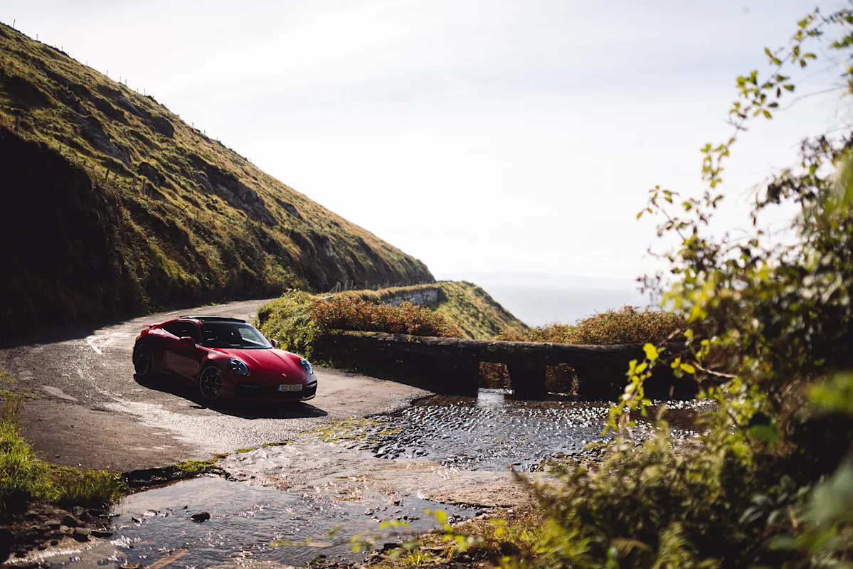 Roter Porsche an Küstenstraße bei Grashügel unter klarem Himmel.